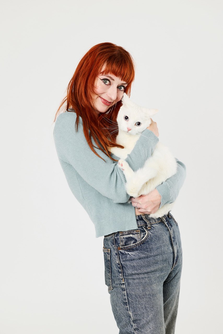 A red-haired woman smiling as she hugs a white cat