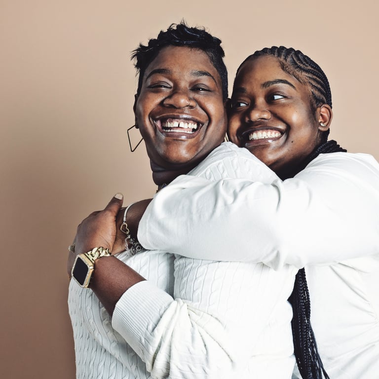 Two women in white tops hugging tightly and laughing on a warm beige backdrop