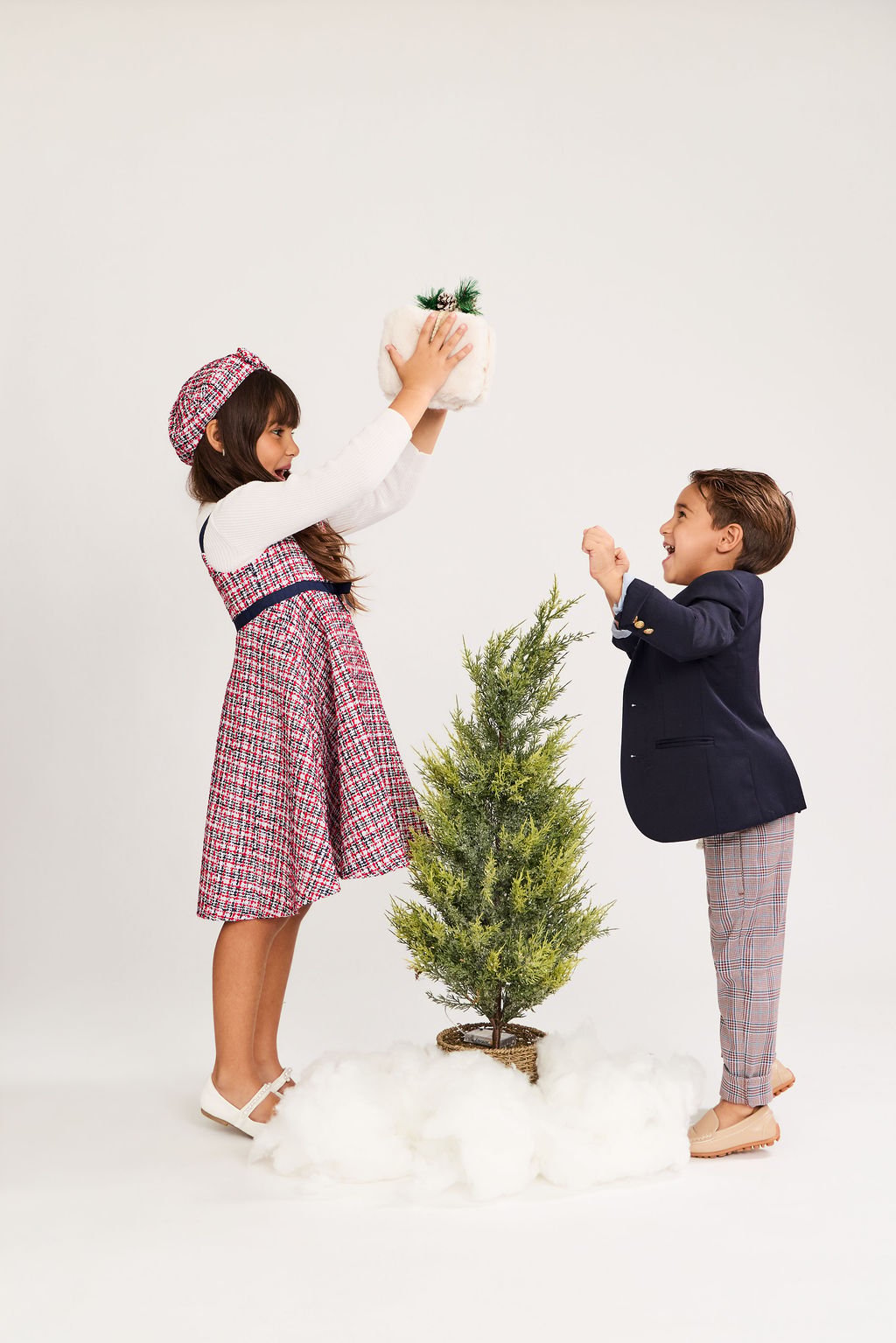 A girl in a tweed dress lifting a tree topper over a little tree in fake snow as her brother reaches up, on a white backdrop