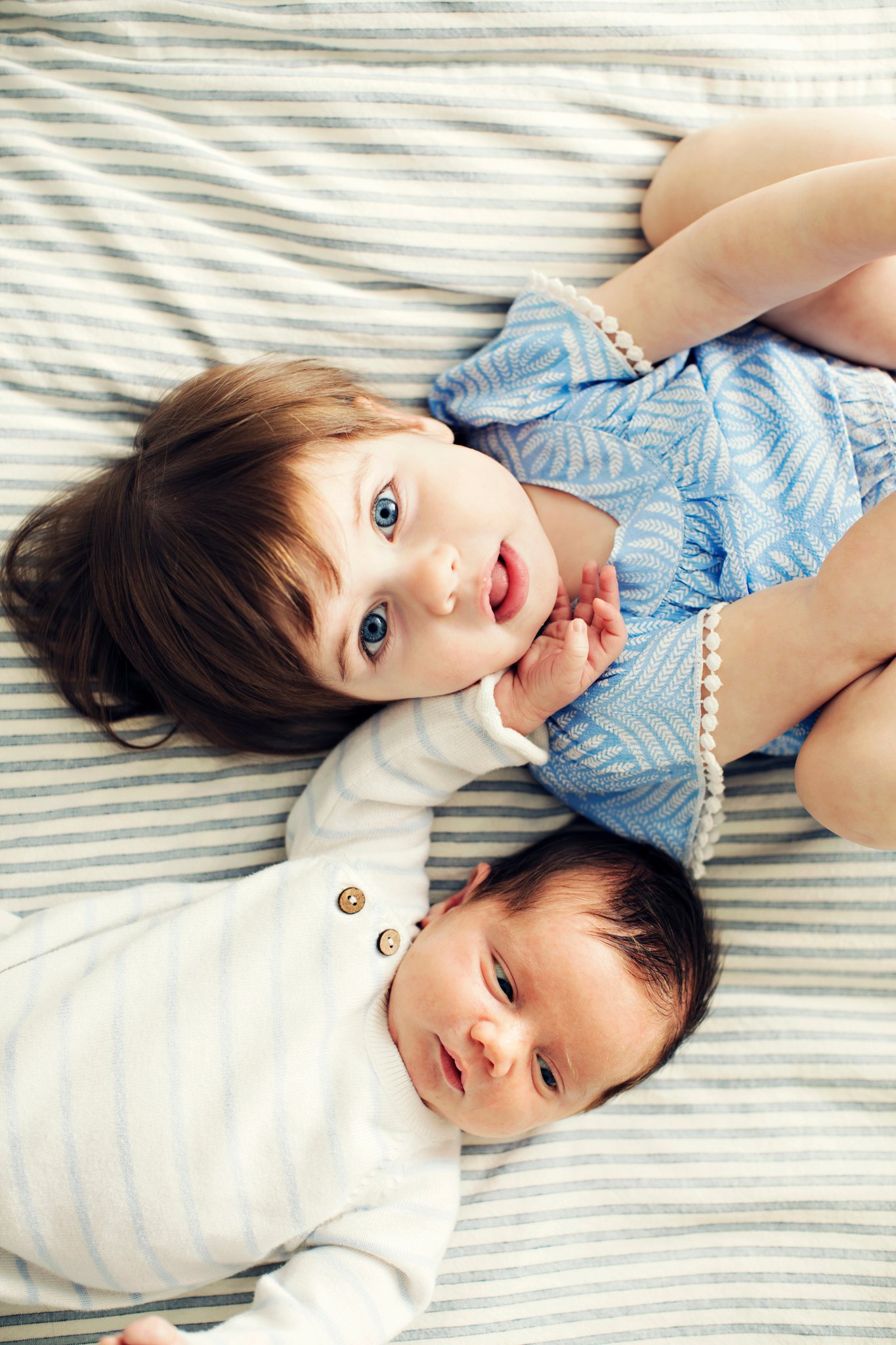 A big sister lying beside her newborn brother on a striped bed