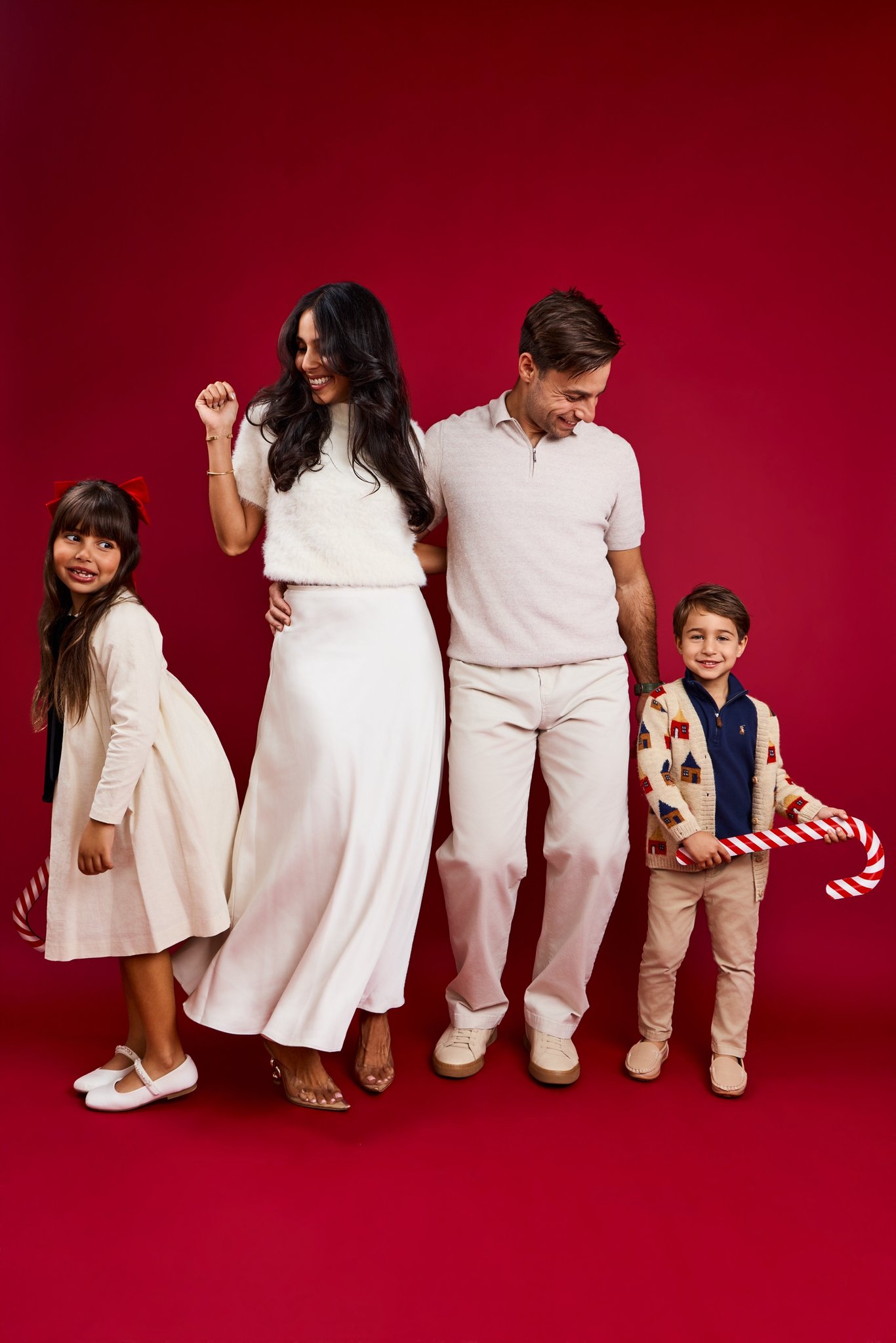 A family of four in cream and white dancing on a red backdrop, the kids holding giant candy canes