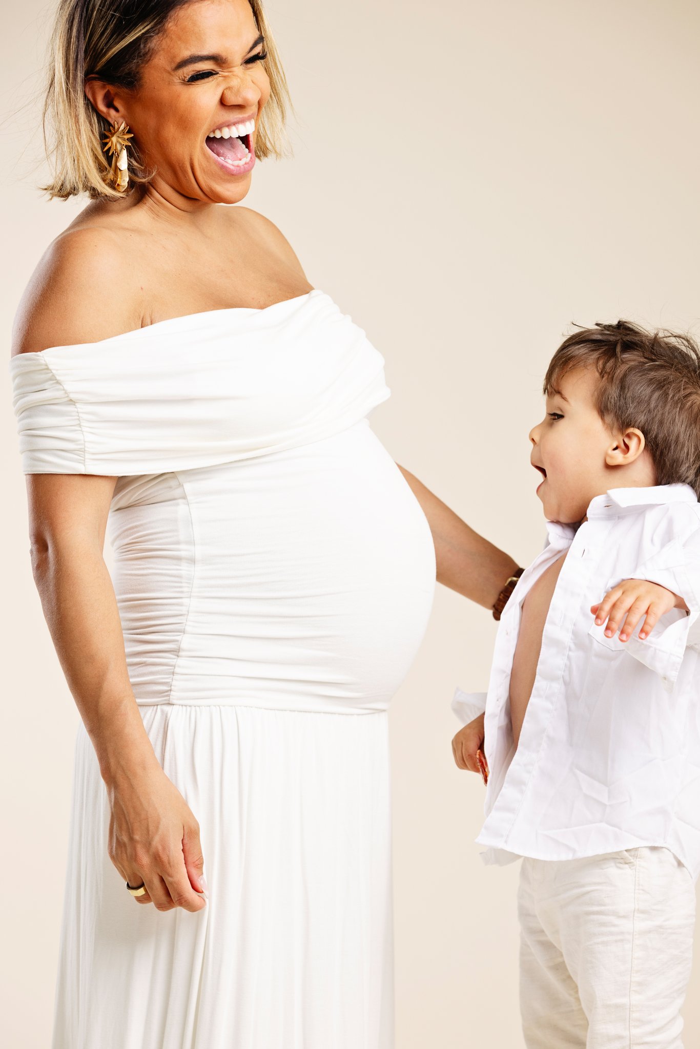 An expectant mother in a white dress laughing, her young son looking up at her bump