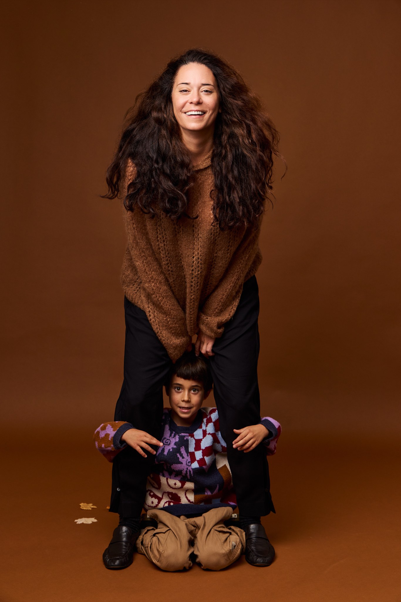 A smiling woman in a brown sweater on the Cinnamon backdrop, a boy peeking out between her legs