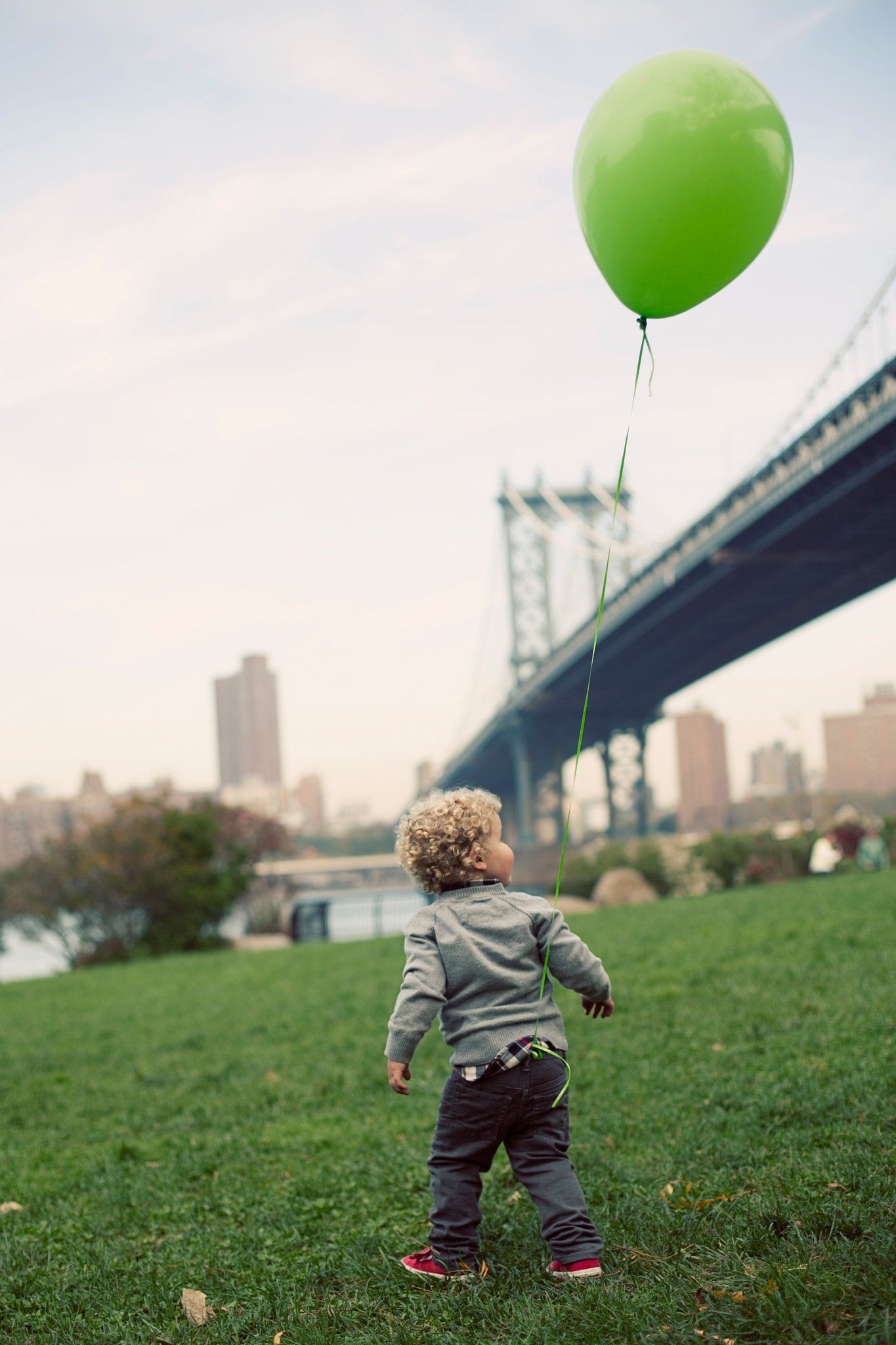 A toddler on the grass watching a green balloon float up beside the Manhattan Bridge