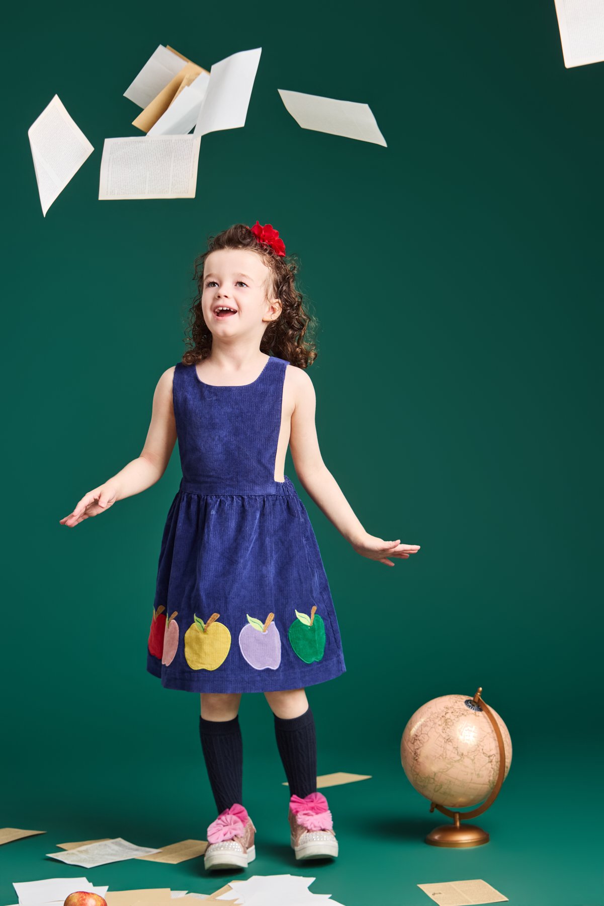 A girl in an apple corduroy dress tossing book pages on the chalkboard-green set