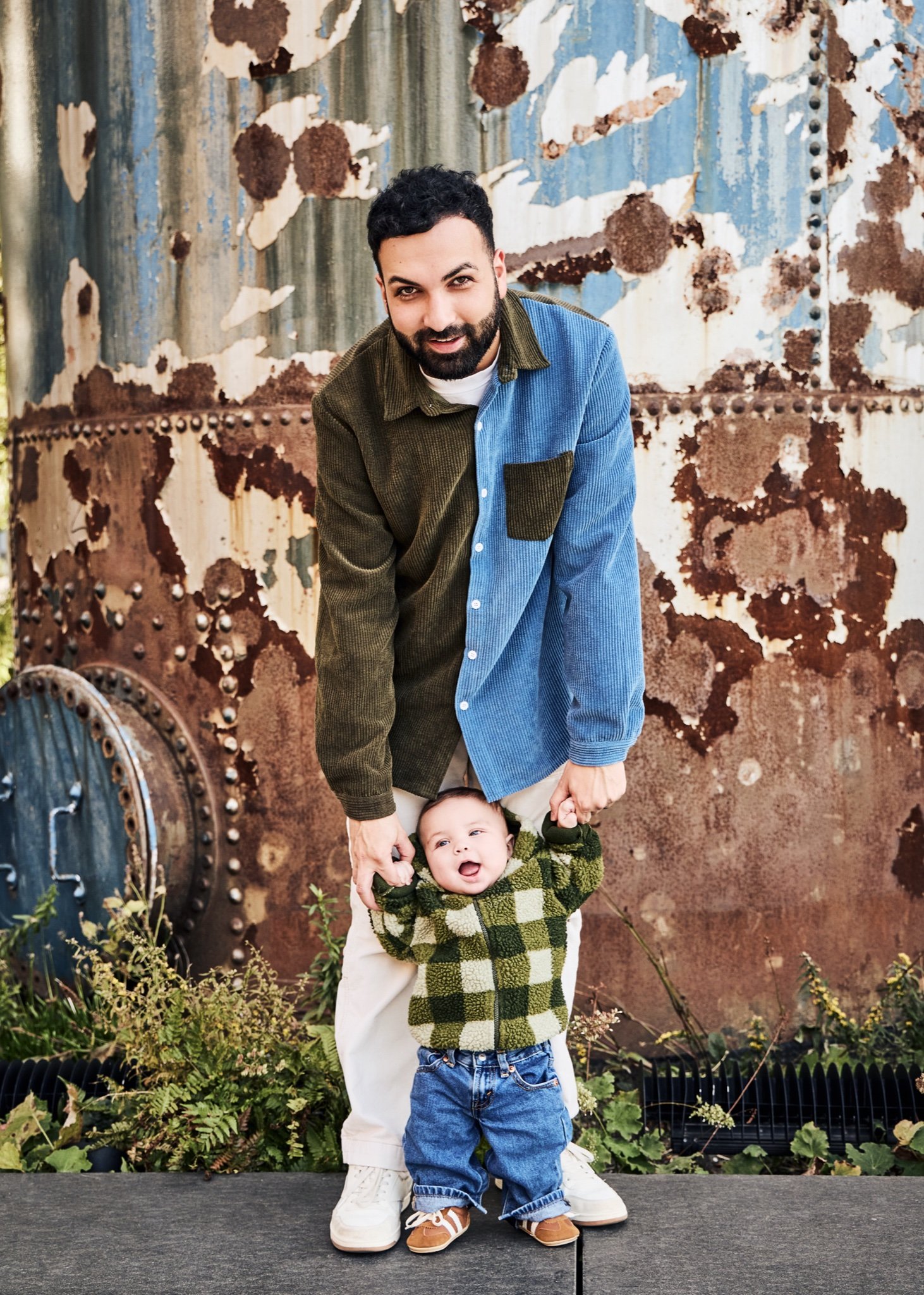 A smiling father helping his baby stand in front of a rusted, paint-peeled steel wall