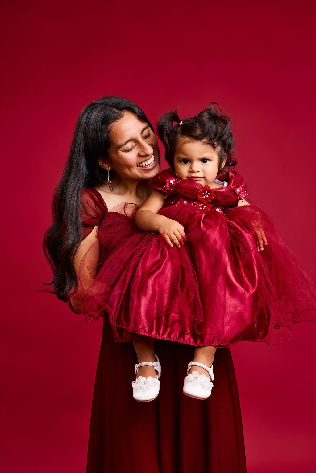 A mother in a deep red gown holding up her smiling baby in a red tulle party dress, on a red backdrop