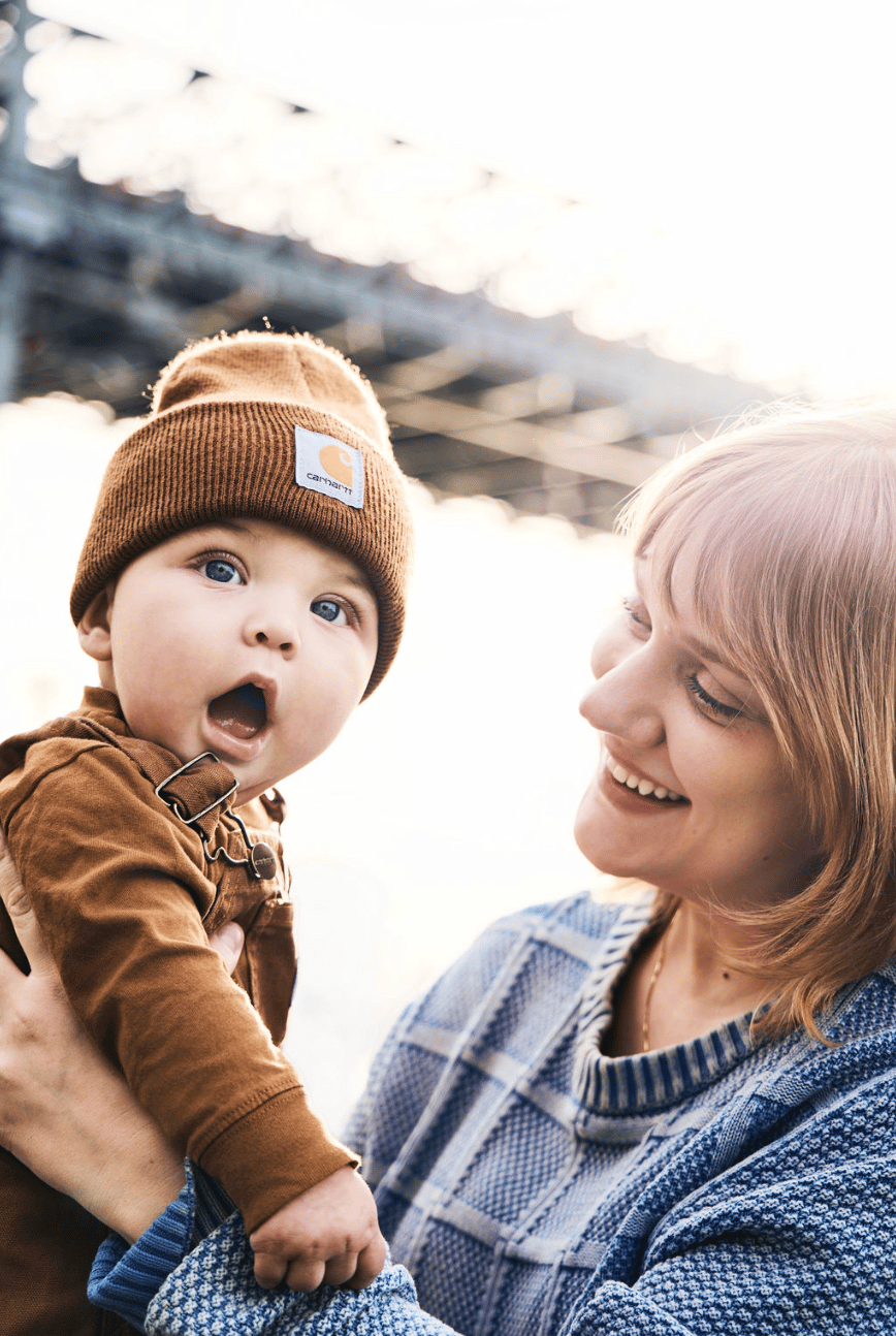 A baby in a brown beanie gasping with delight as his mother smiles at him