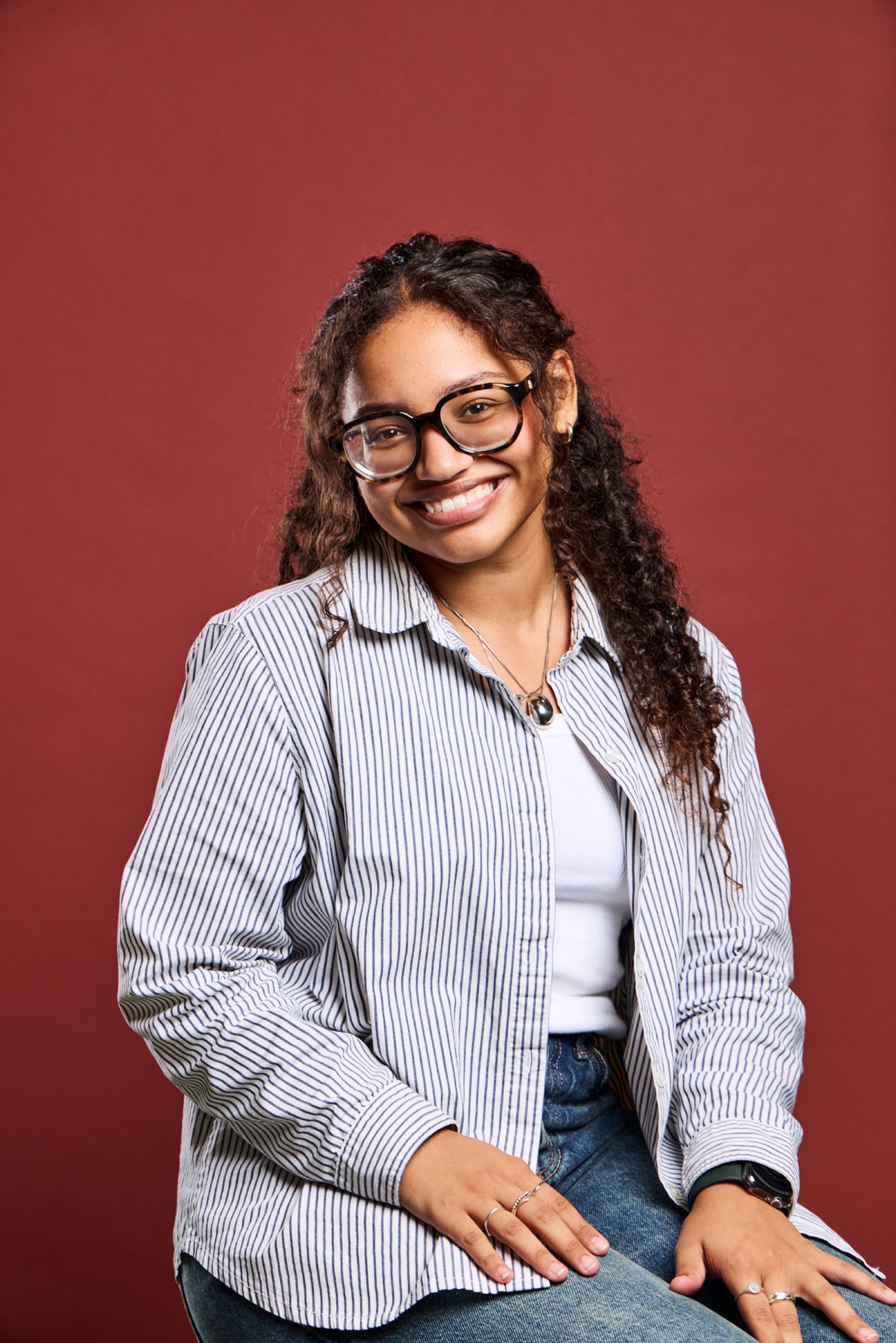Penelope, Daysee photographer, smiling in glasses and a striped shirt on a red backdrop