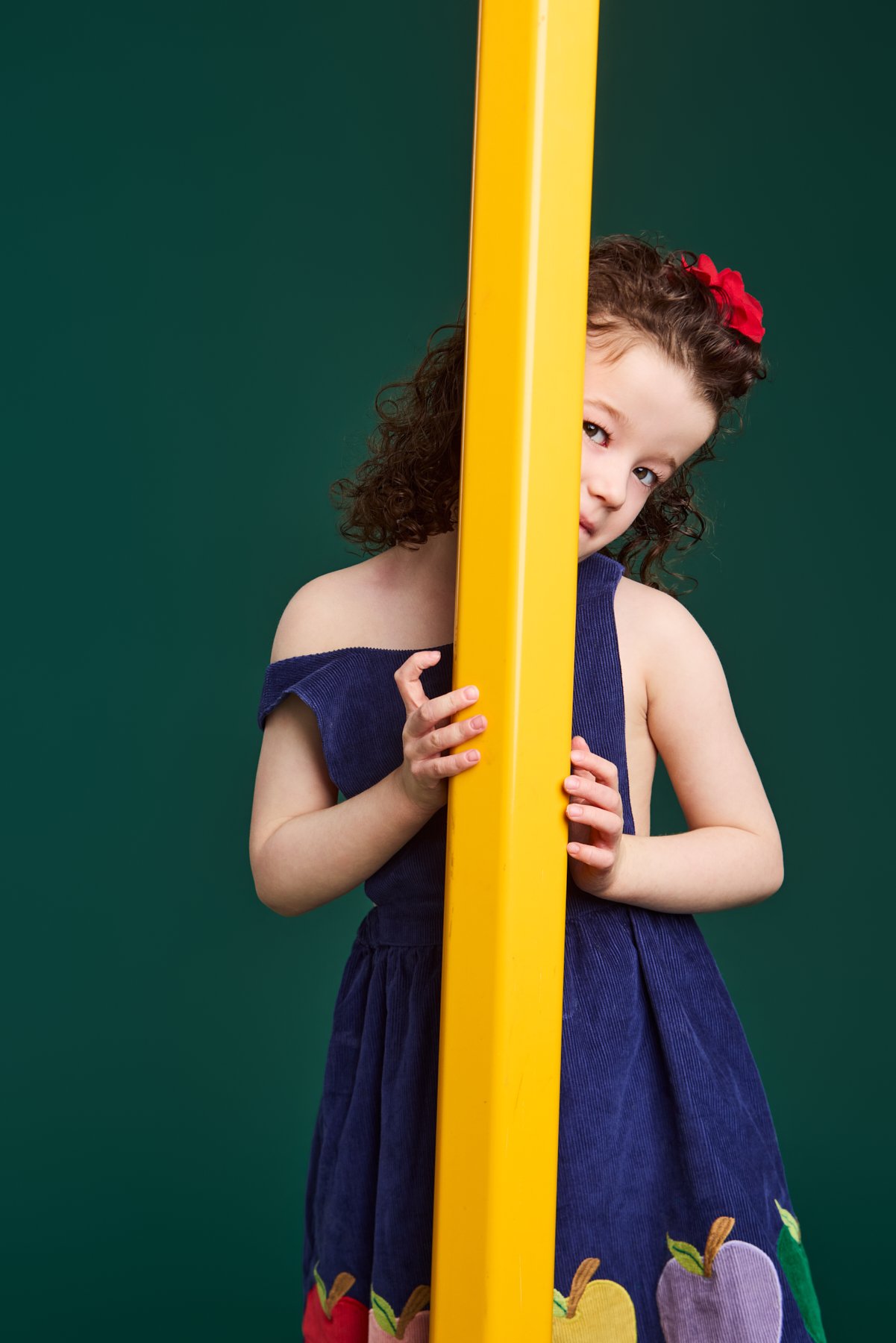 A girl in an apple dress peeking out from behind a giant yellow pencil