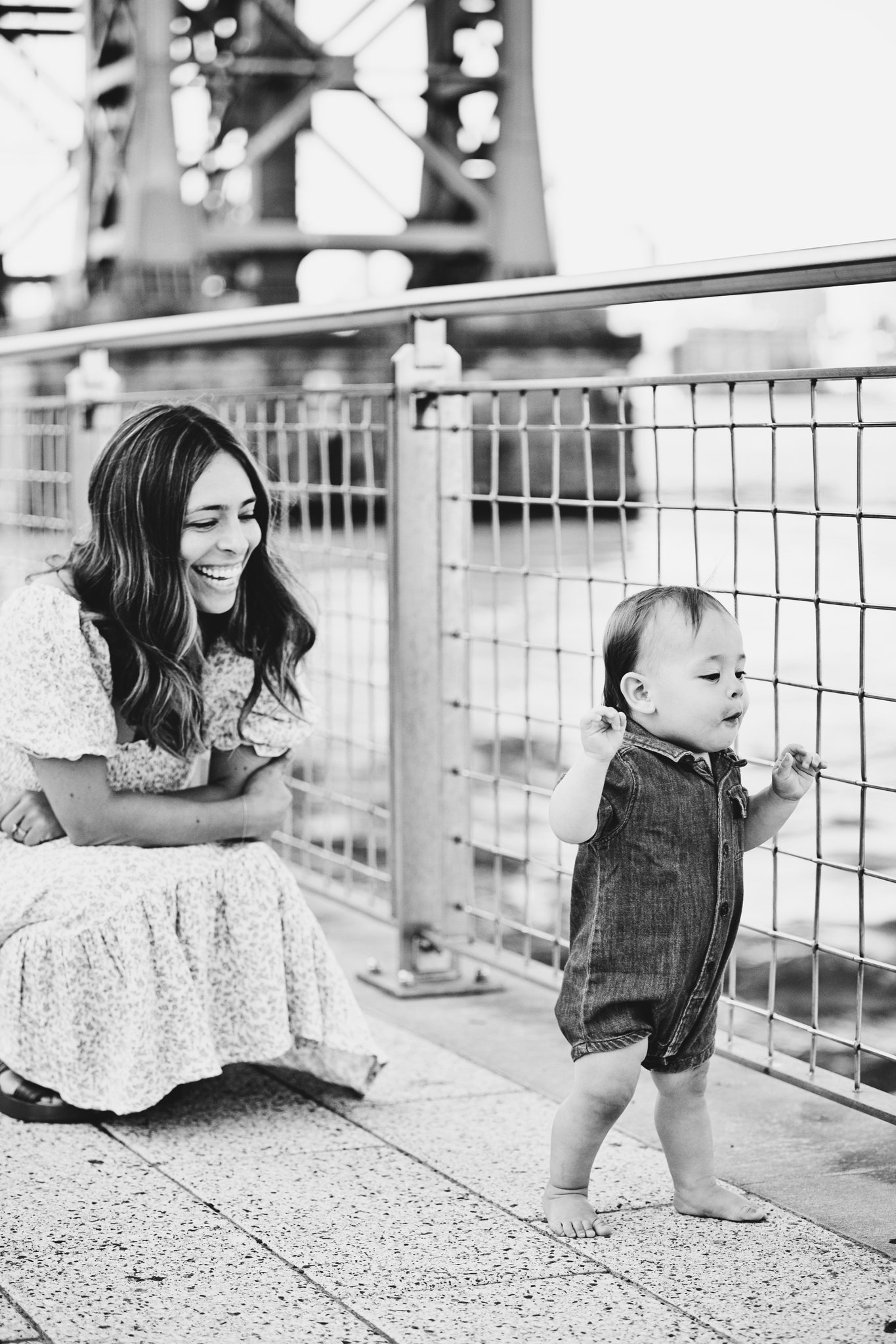 A mother crouching by the riverside railing, smiling at her toddler, in black and white