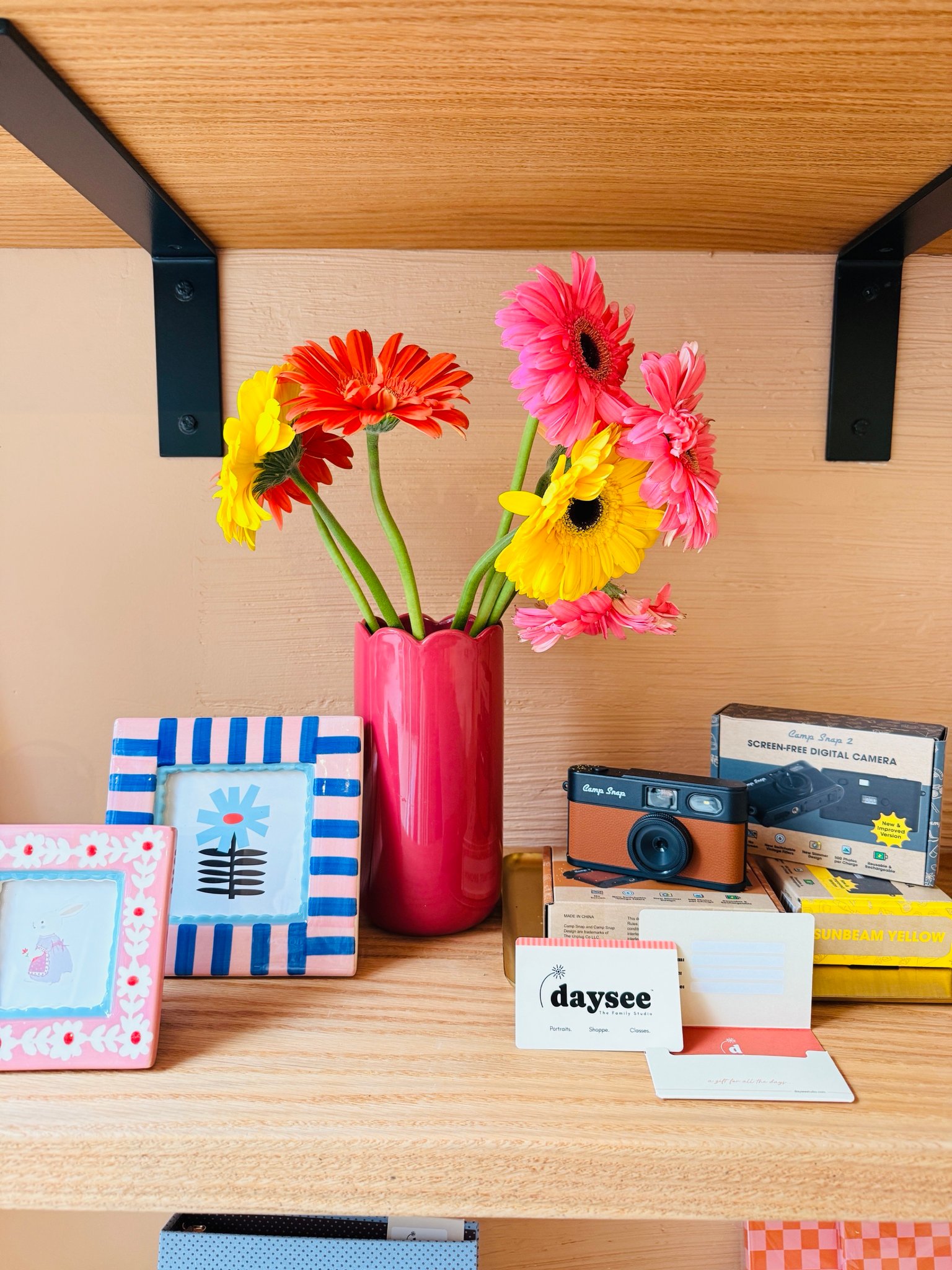 A Daysee gift card on a shop shelf beside gerbera daisies, painted frames and a Camp Snap camera