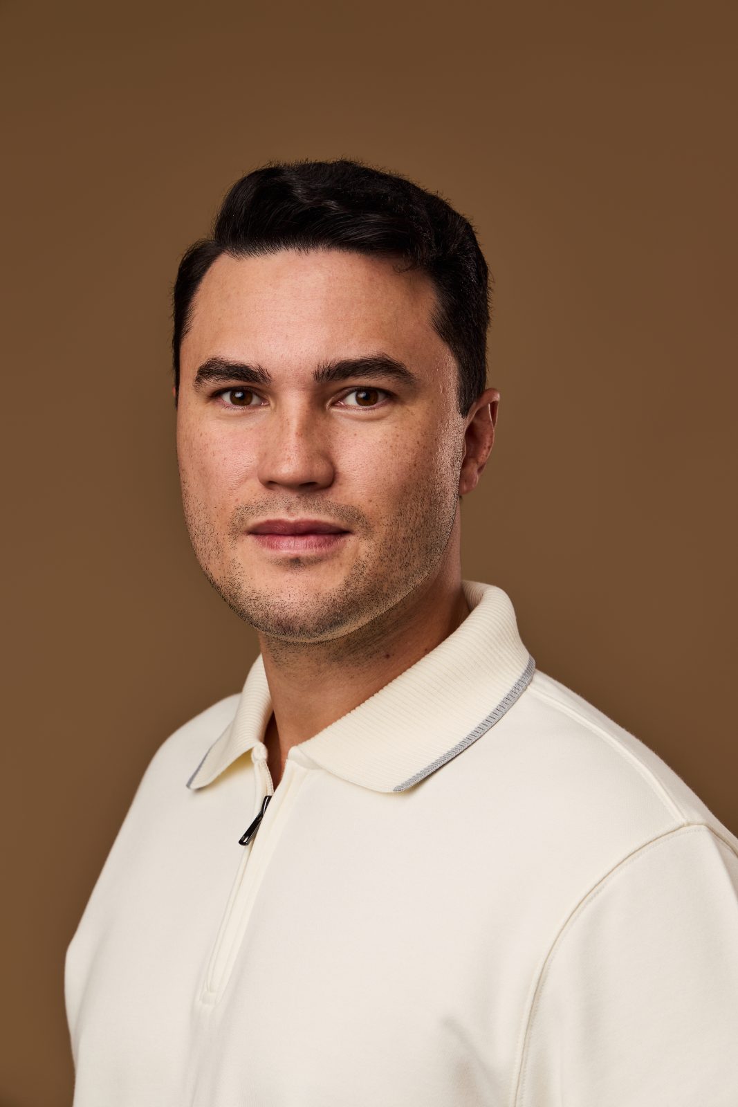 Headshot of a man in a cream polo on a brown backdrop