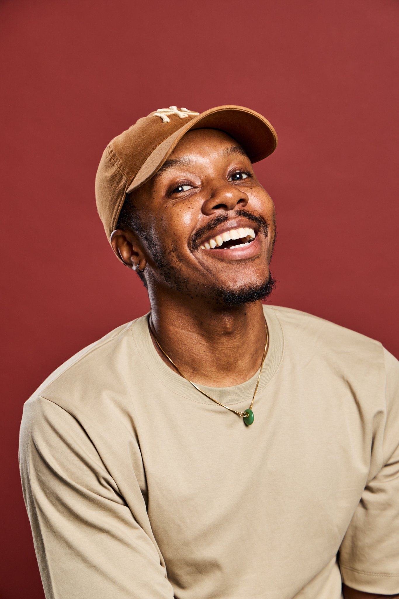Neriah, Daysee photographer, laughing in a tan cap and t-shirt on a red backdrop