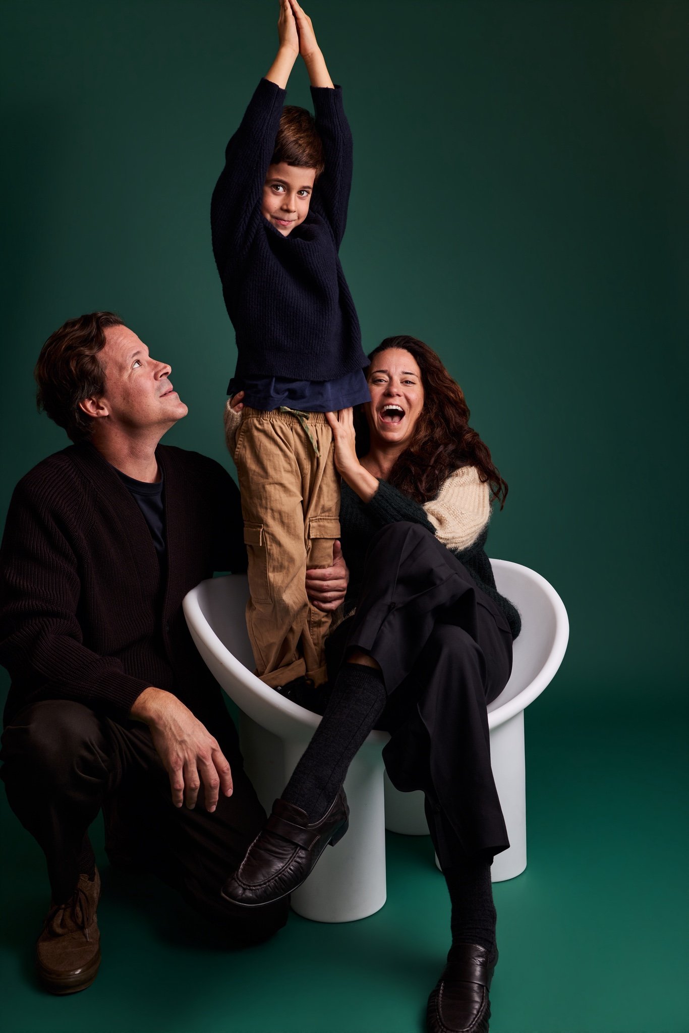 A boy standing on a white chair with his arms stretched overhead as his parents look up and laugh, on the Evergreen backdrop