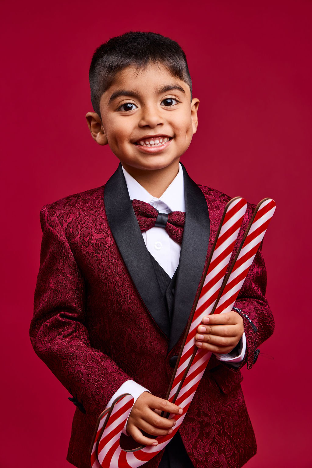 A smiling boy in a burgundy brocade tuxedo jacket and bow tie holding two giant candy canes, on a red backdrop
