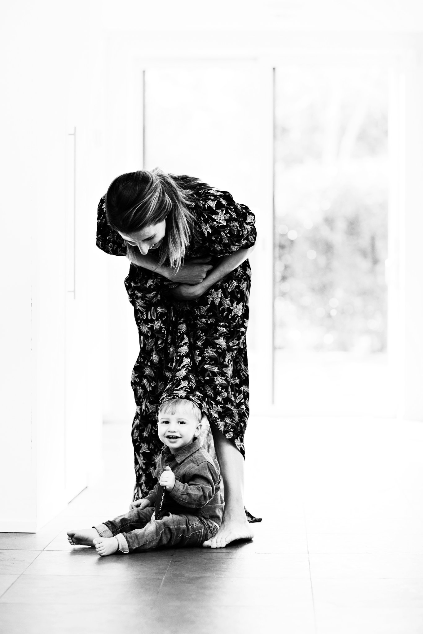 A mother leaning over her laughing toddler in a bright doorway, in black and white