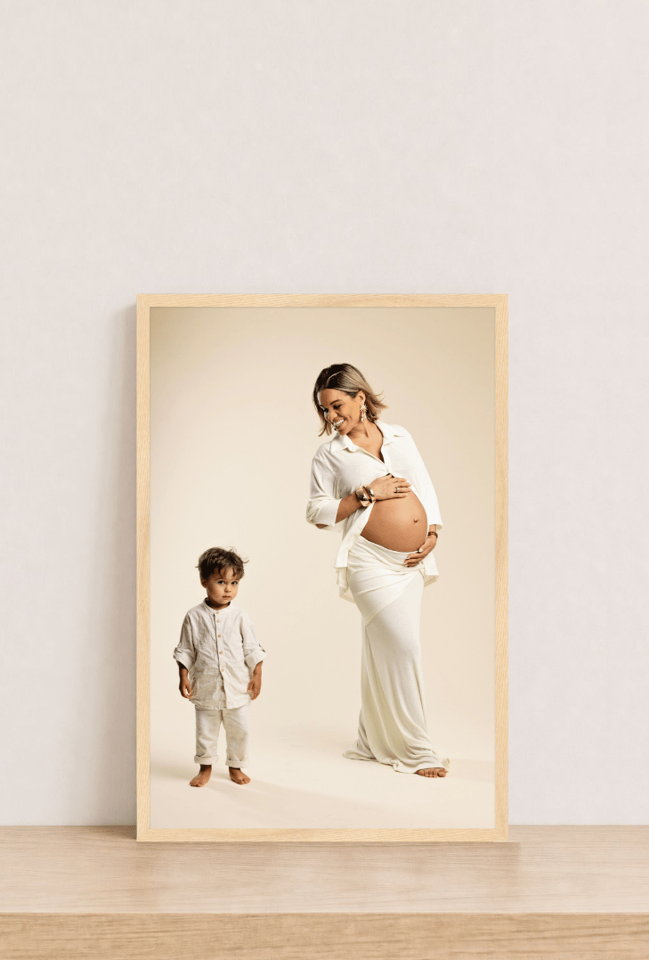 A light oak frame holding a portrait of a mother cradling her bump beside her toddler, leaning on a wood shelf