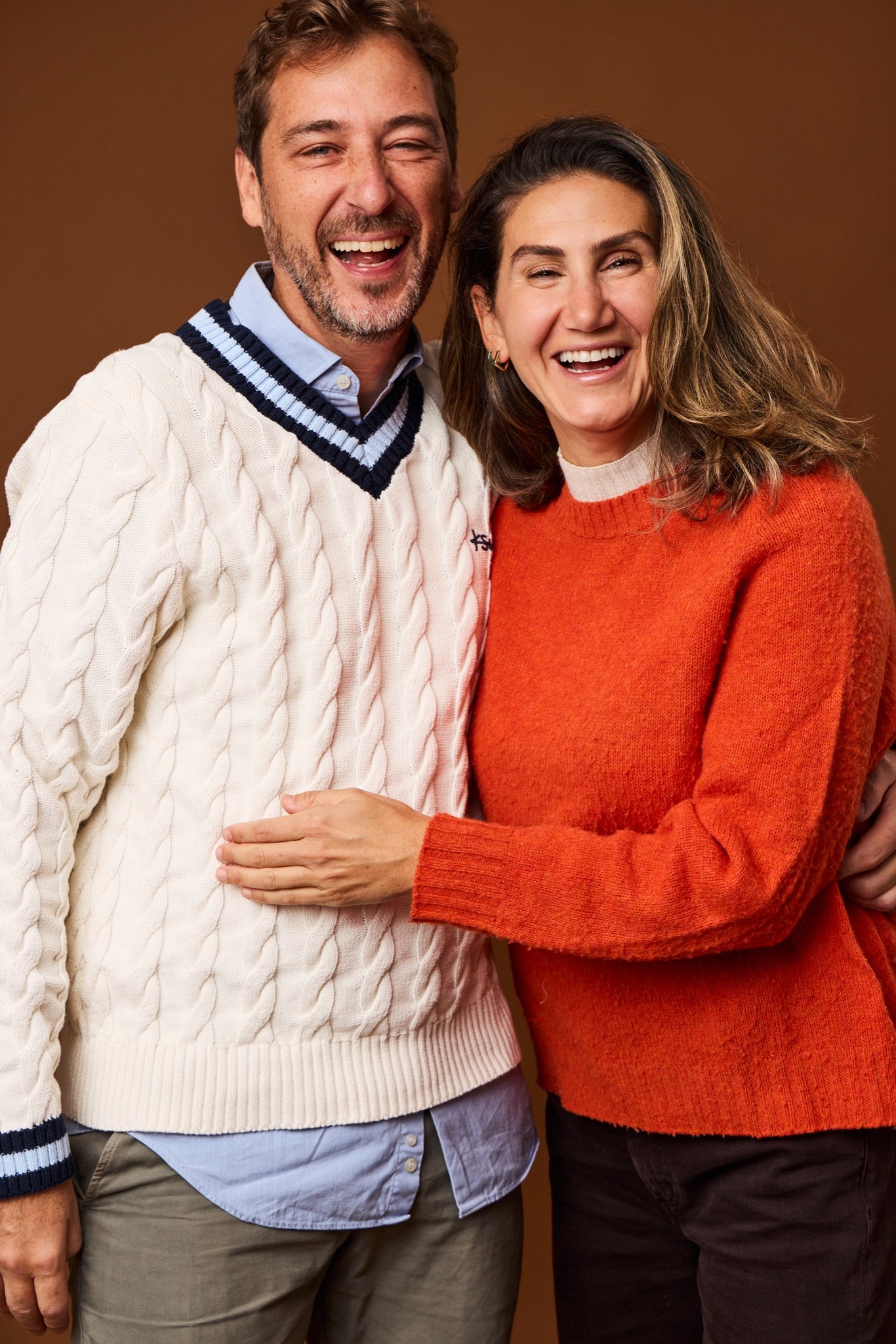A laughing couple arm in arm in a cream cable-knit sweater and an orange sweater, on the Cinnamon backdrop