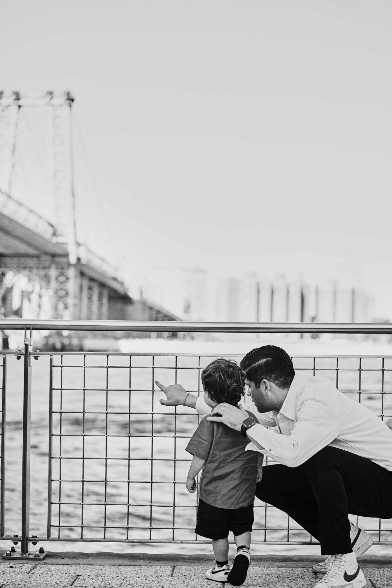 A father crouching with his son at the riverside railing, in black and white