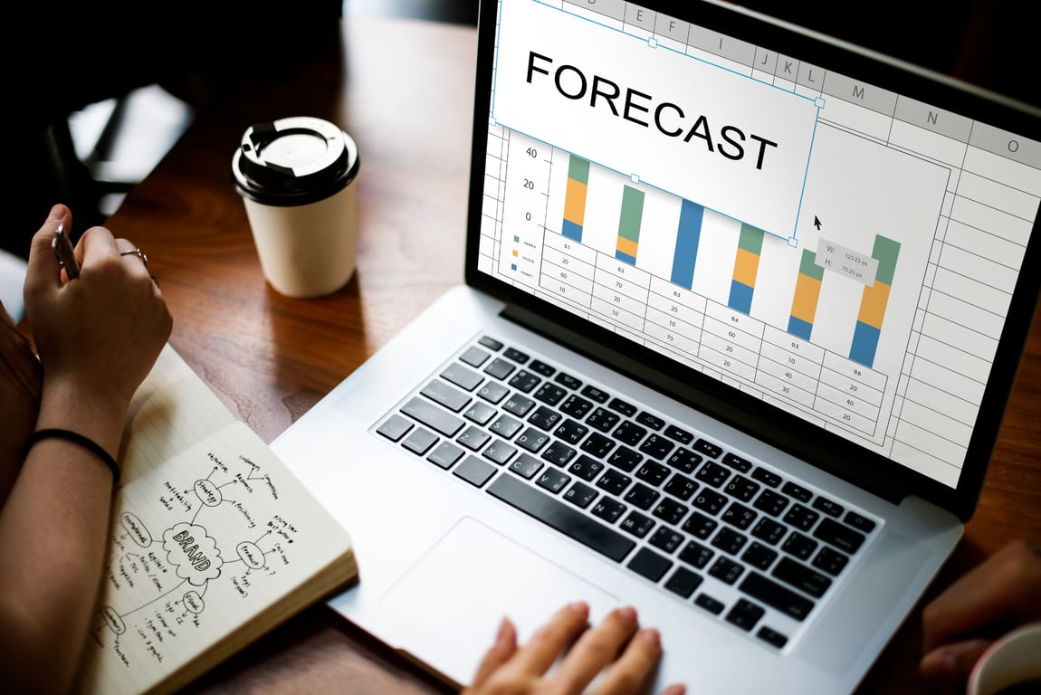 A hand working on a laptop showing a Forecast Data sheet for predicting future demand in the hospitality industry.