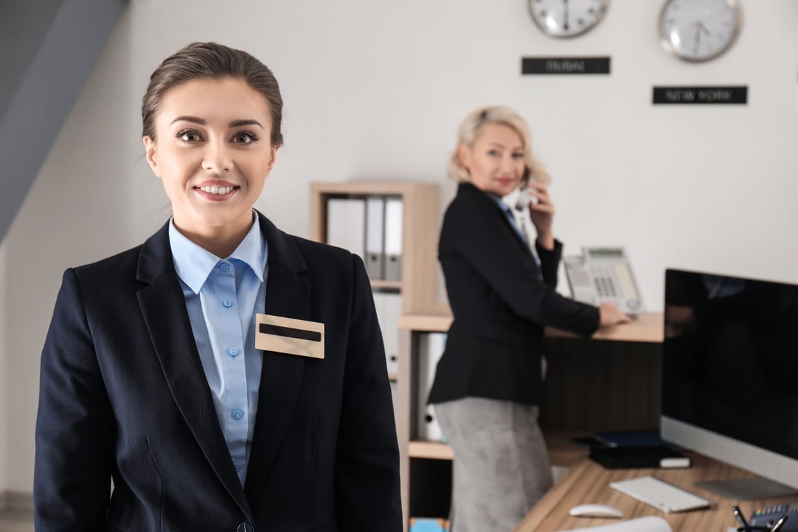 Smiling female hotel receptionist wearing uniform, with businesswoman on phone in background