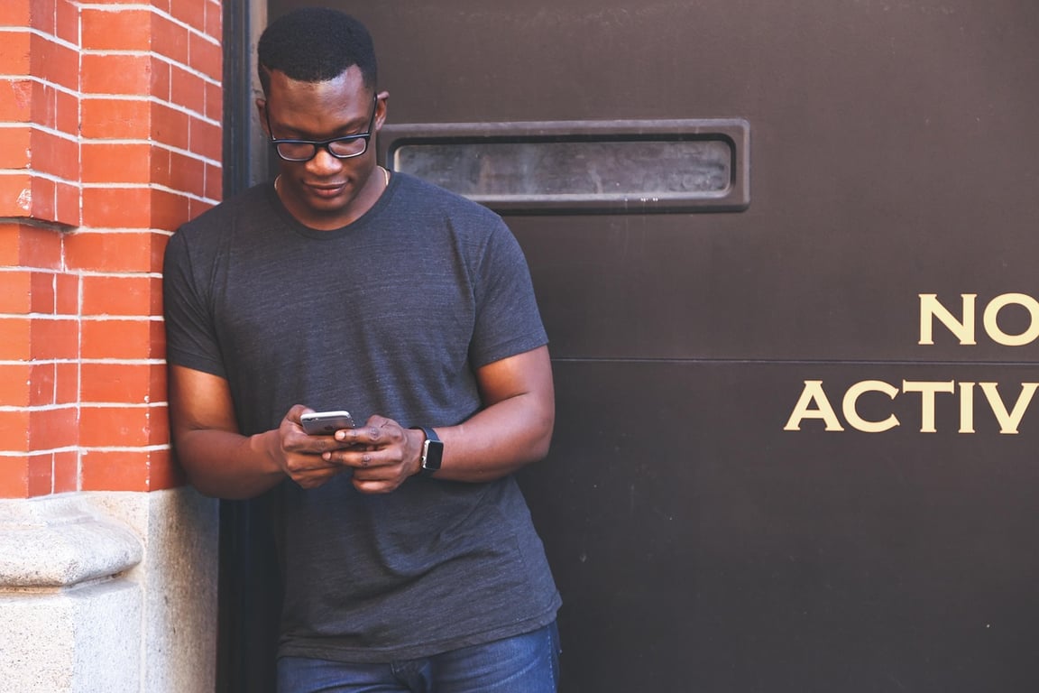 A man wearing spectacles standing by a door using his mobile phone.