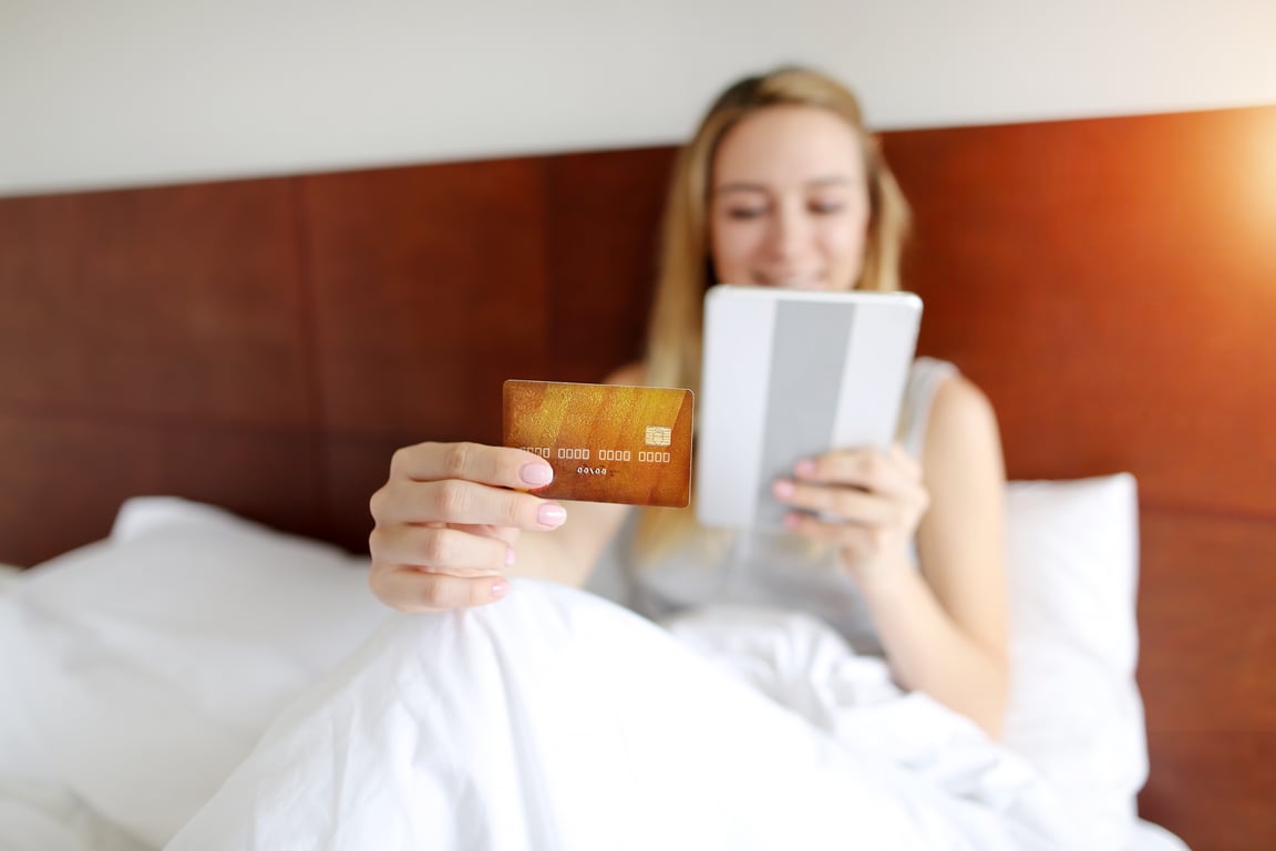 Woman in hotel bed holding credit card and tablet device for online payment processing