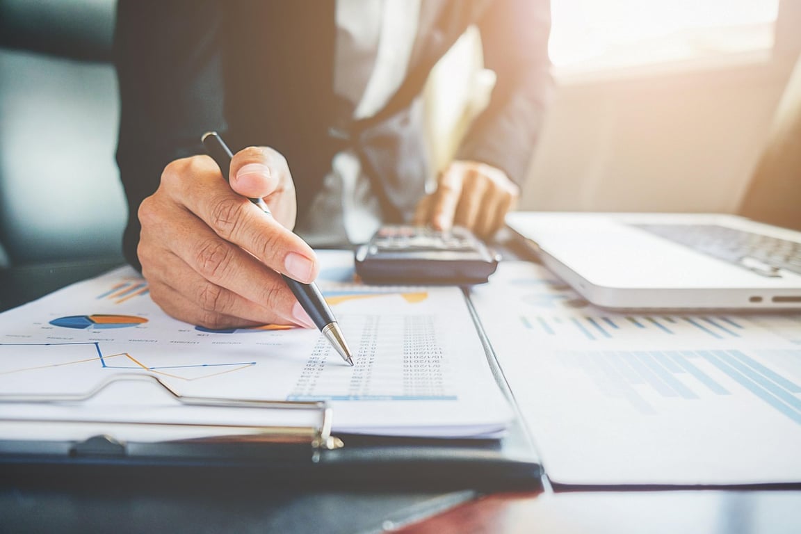 A professional analyzing printed charts with a pen, calculator, and laptop, representing hands-on approach towards hotel demand forecasting.