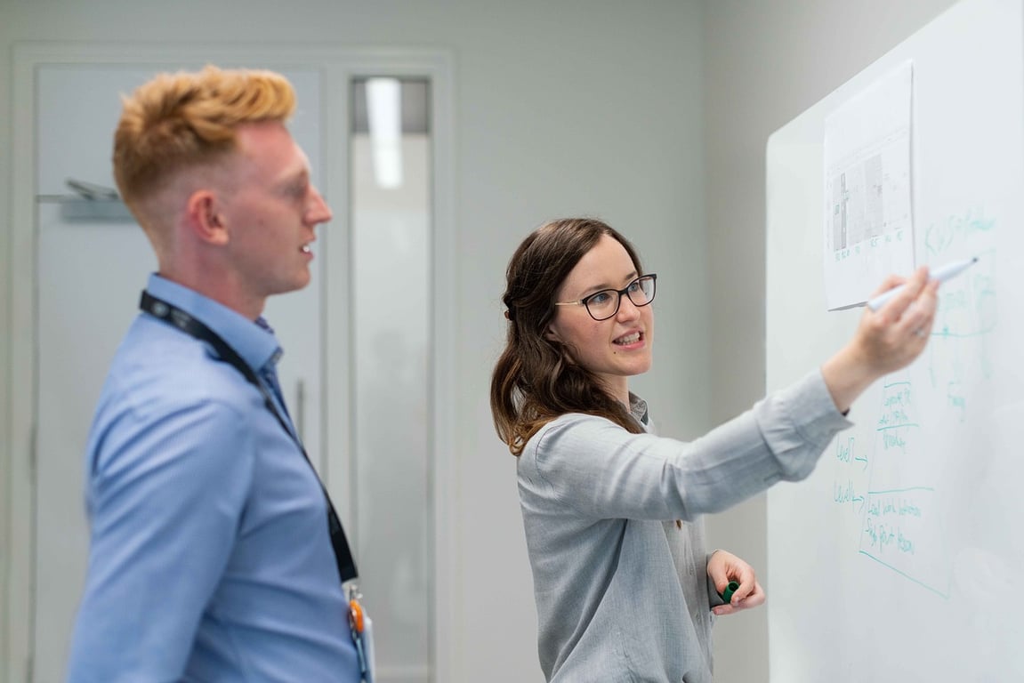 Two business professionals working, one is pointing to a digital strategy diagram on a clear whiteboard, representing brand strategy planning.