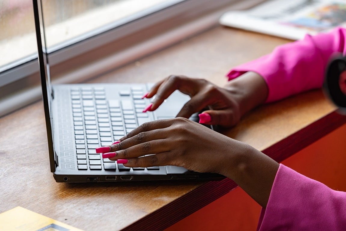 A woman with well-manicured nails working on a laptop that’s placed on a window ledge.