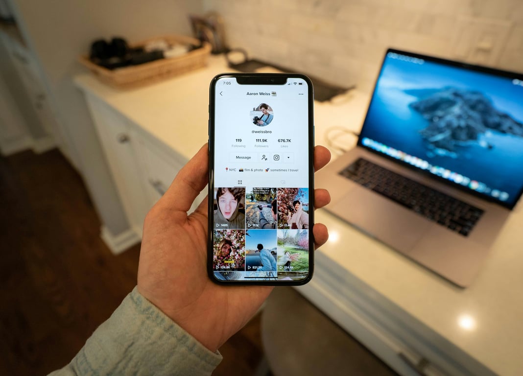 Hand holding smartphone showing TikTok profile with travel-related videos on laptop in background
