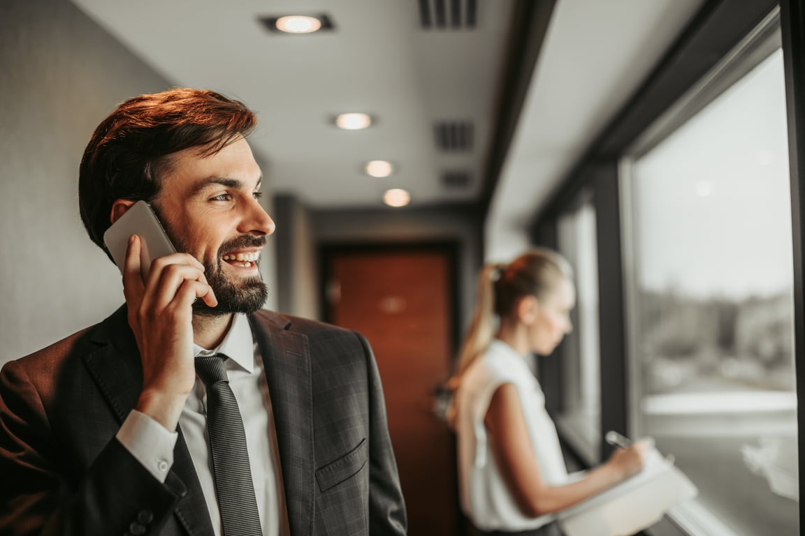 Smiling businessman on phone in office hallway, with businesswoman in background