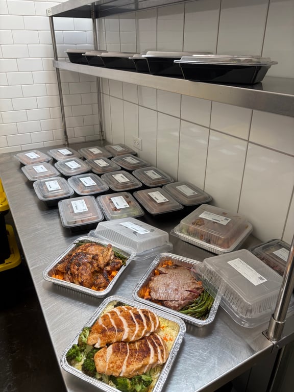 A stainless steel prep table in an Austin commissary kitchen covered in labeled Full Time Food meal containers, with family trays of roasted chicken, sliced flank steak, and chicken breast over rice in front