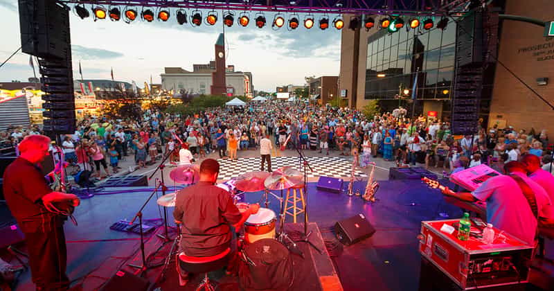 On-stage view from behind the drummer looking out at a large crowd in downtown Elkhart at dusk during the Elkhart Jazz Festival, with the historic clock tower behind the buildings.
