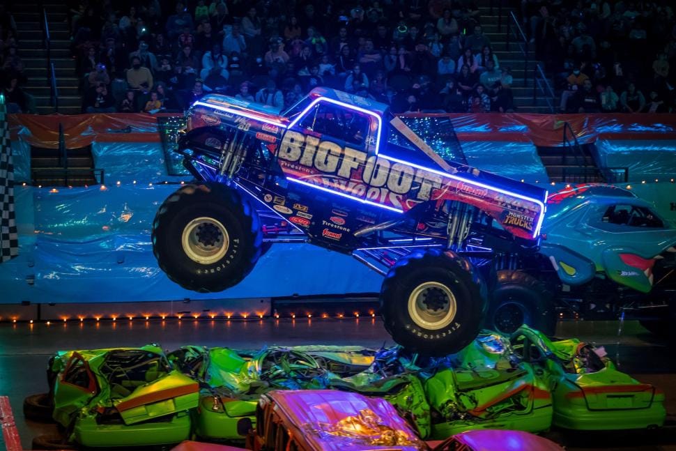 The Bigfoot monster truck outlined in glowing blue neon, jumping over a row of crushed cars at a Hot Wheels Monster Trucks Live arena show