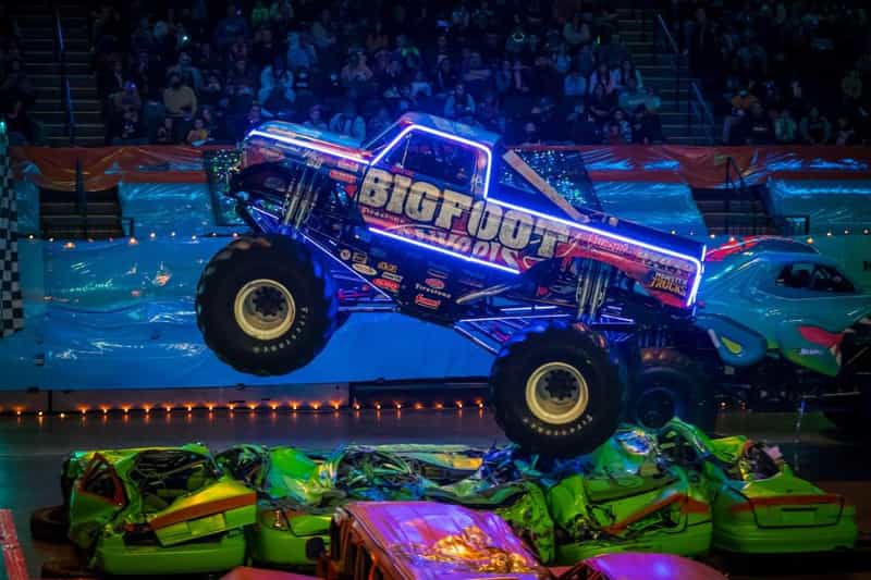 The Bigfoot monster truck outlined in glowing blue neon, jumping over a row of crushed cars at a Hot Wheels Monster Trucks Live arena show