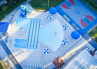 Aerial view of the Blue Devil Lagoon community pool in Kentland, Indiana, with a blue waterslide, lap lanes, shade umbrellas and basketball courts next to the deck