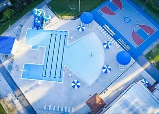 Aerial view of the Blue Devil Lagoon community pool in Kentland, Indiana, with a blue waterslide, lap lanes, shade umbrellas and basketball courts next to the deck