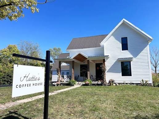 The white farmhouse exterior of Hattie's Coffee House in Whitestown, Indiana, with a black-and-white yard sign out front and rocking chairs on the covered porch