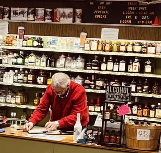 A man in a red sweater working behind the counter at Holly's Package Store in Greentown, Indiana, with shelves of liquor bottles and a hand-lettered store hours sign above him