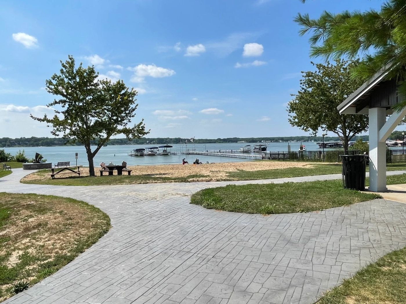Sandy public beach on Cedar Lake, Indiana, with a fishing pier, pontoon boats and a stamped-concrete lakefront park path under a blue summer sky