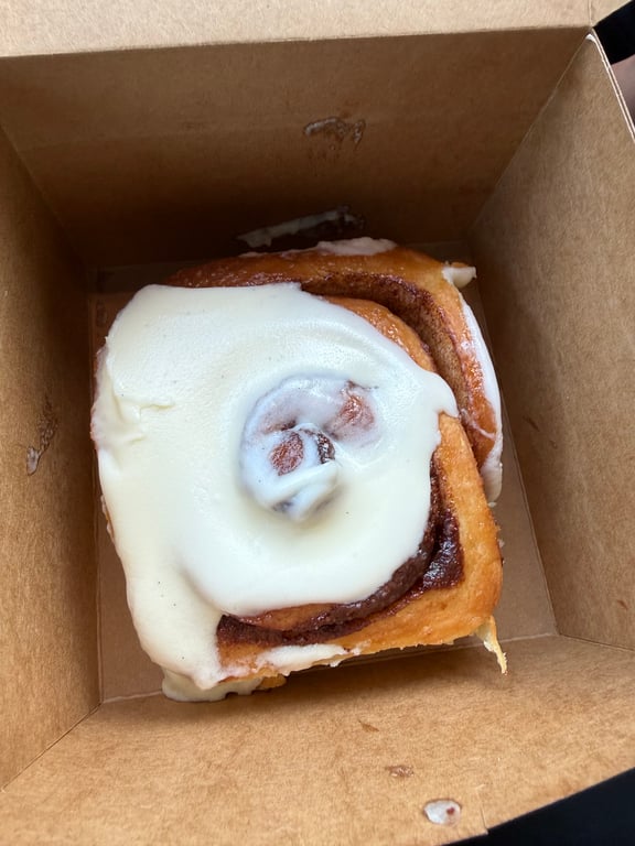 A large square cinnamon roll topped with thick cream cheese frosting in a kraft takeout box from René's Bakery in Broad Ripple.