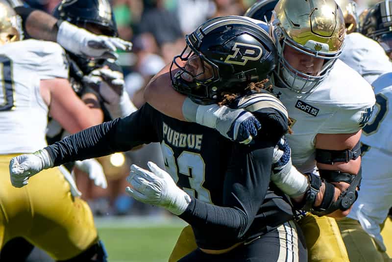A Notre Dame lineman in a white jersey and gold helmet grabs a Purdue pass rusher in a black jersey and helmet with the gold P logo at Ross-Ade Stadium
