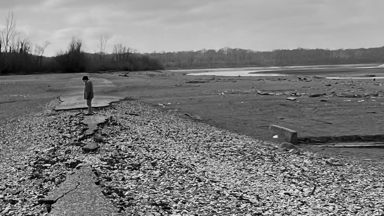 A lone person stands on a cracked, exposed roadbed in the drained basin of Mississinewa Lake near Somerset, Indiana, under an overcast winter sky.