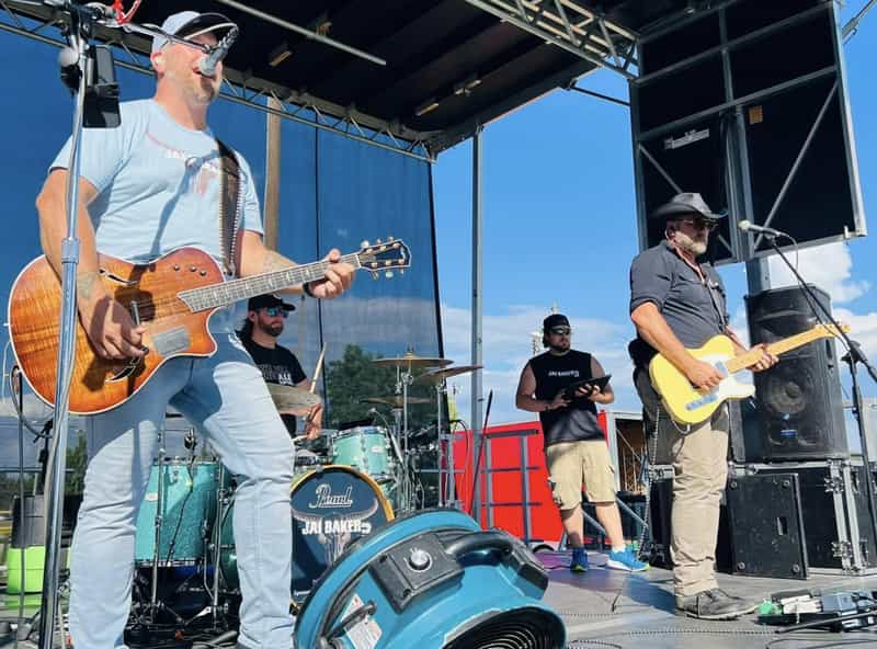 A country rock band performs on an outdoor stage under blue skies at WAMMfest in Greenwood, with a singer on guitar and a bandmate in a cowboy hat