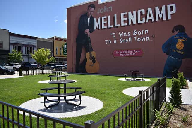 The John Mellencamp mural on a brick building in downtown Seymour, Indiana, reading “I Was Born In A Small Town,” with picnic tables on the lawn of the adjacent pocket park