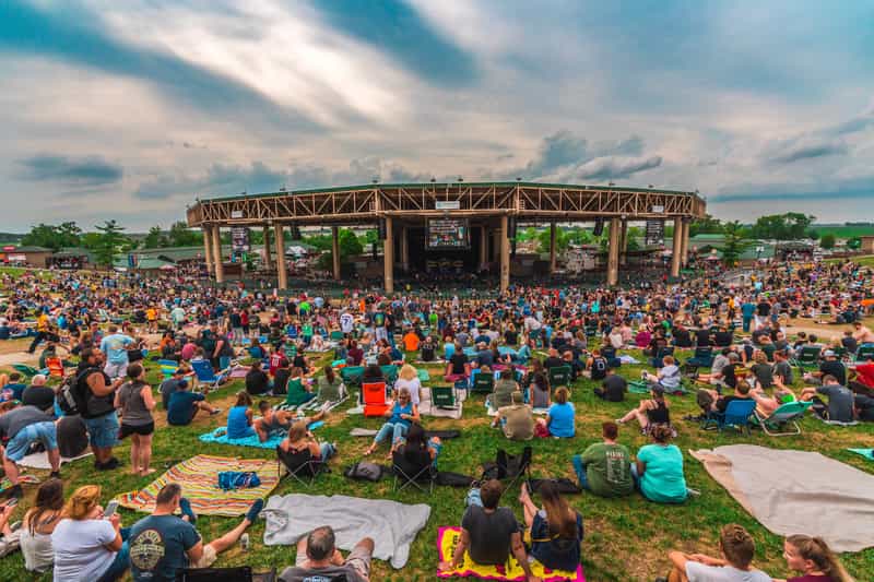 A large crowd fills the lawn at Ruoff Music Center in Noblesville, Indiana, watching a summer concert under a dramatic cloudy sky.