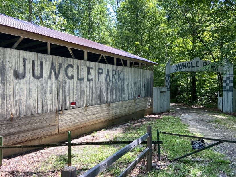 Weathered wooden building with JUNGLE PARK painted on its side beside a wooded gravel drive and a checkered-pattern entrance arch, with a private property no trespassing sign in the foreground at Jungle Park Speedway near Bloomingdale, Indiana