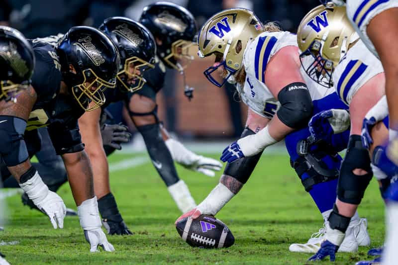 Purdue defensive linemen in black helmets line up across from Washington's center as he grips the ball before the snap at Ross-Ade Stadium